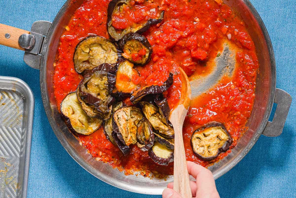 Slices of roasted eggplant being stirred into the pasta all norma sauce in a skillet with a wooden spoon.