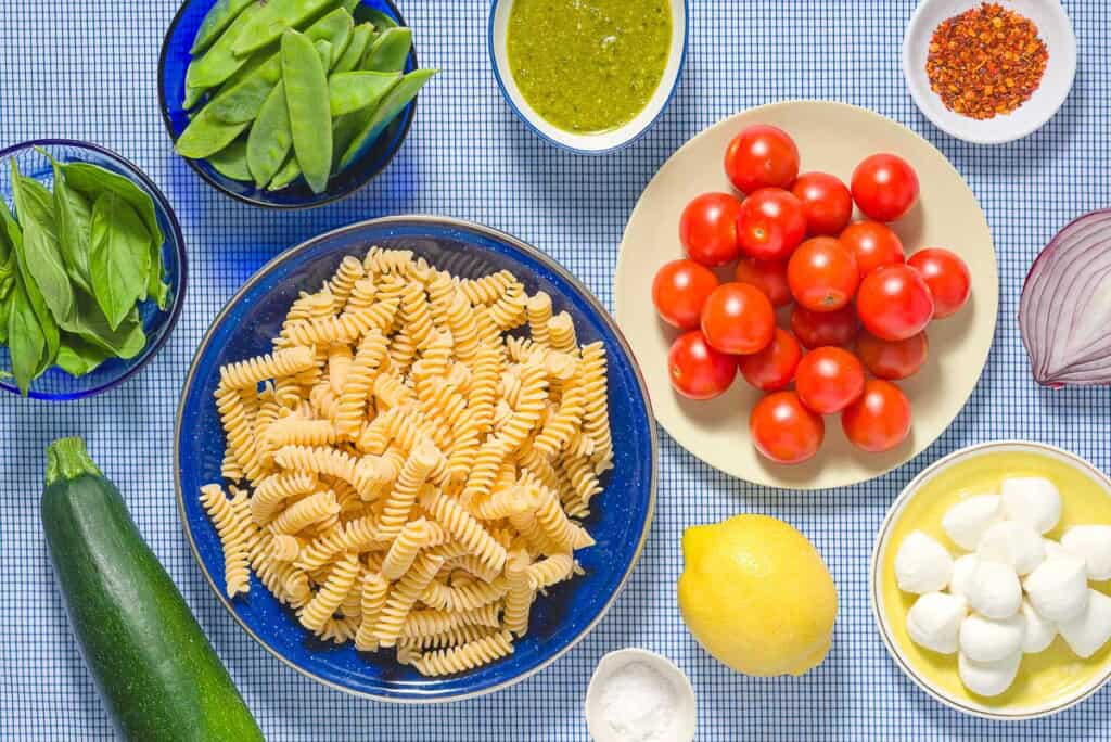 Ingredients for pesto pasta salad including fusilli pasta, salt, zucchini, snap peas, red onions, cherry tomatoes, basil pesto, small mozzarella balls, red pepper flakes, lemon and basil.