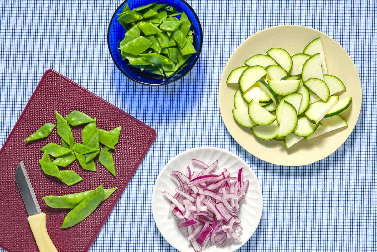Bowls of sliced snap peas, zucchini and red onion next to a cutting board with more sliced snap peas and a knife.