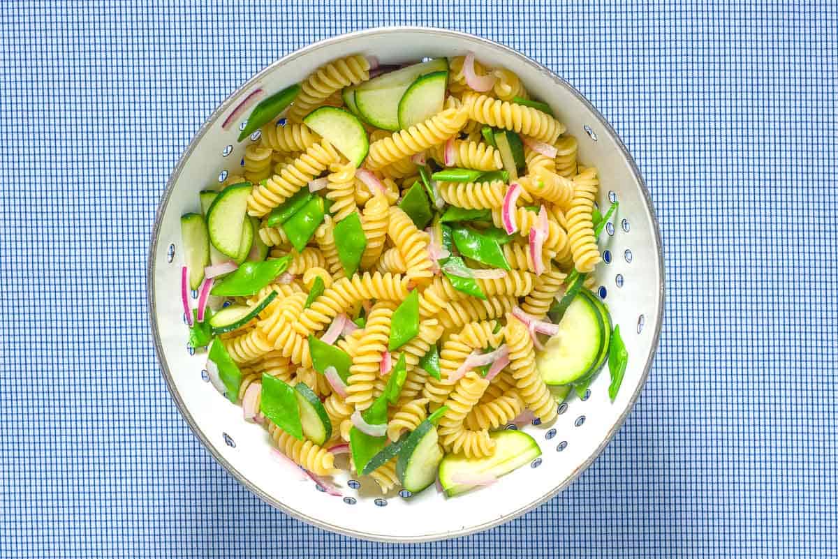 The fusilli for the pasta salad mixed with sliced snap peas, red onion and zucchini in a colander.