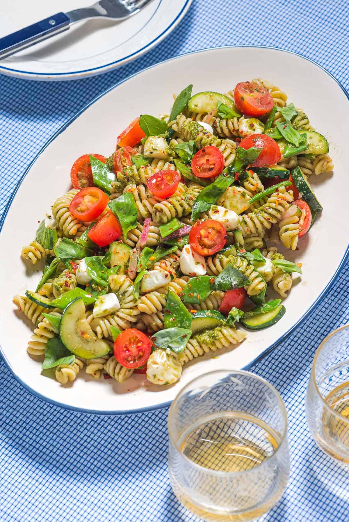 Pesto pasta salad on a platter next to a plate with a fork and two glasses of white wine.