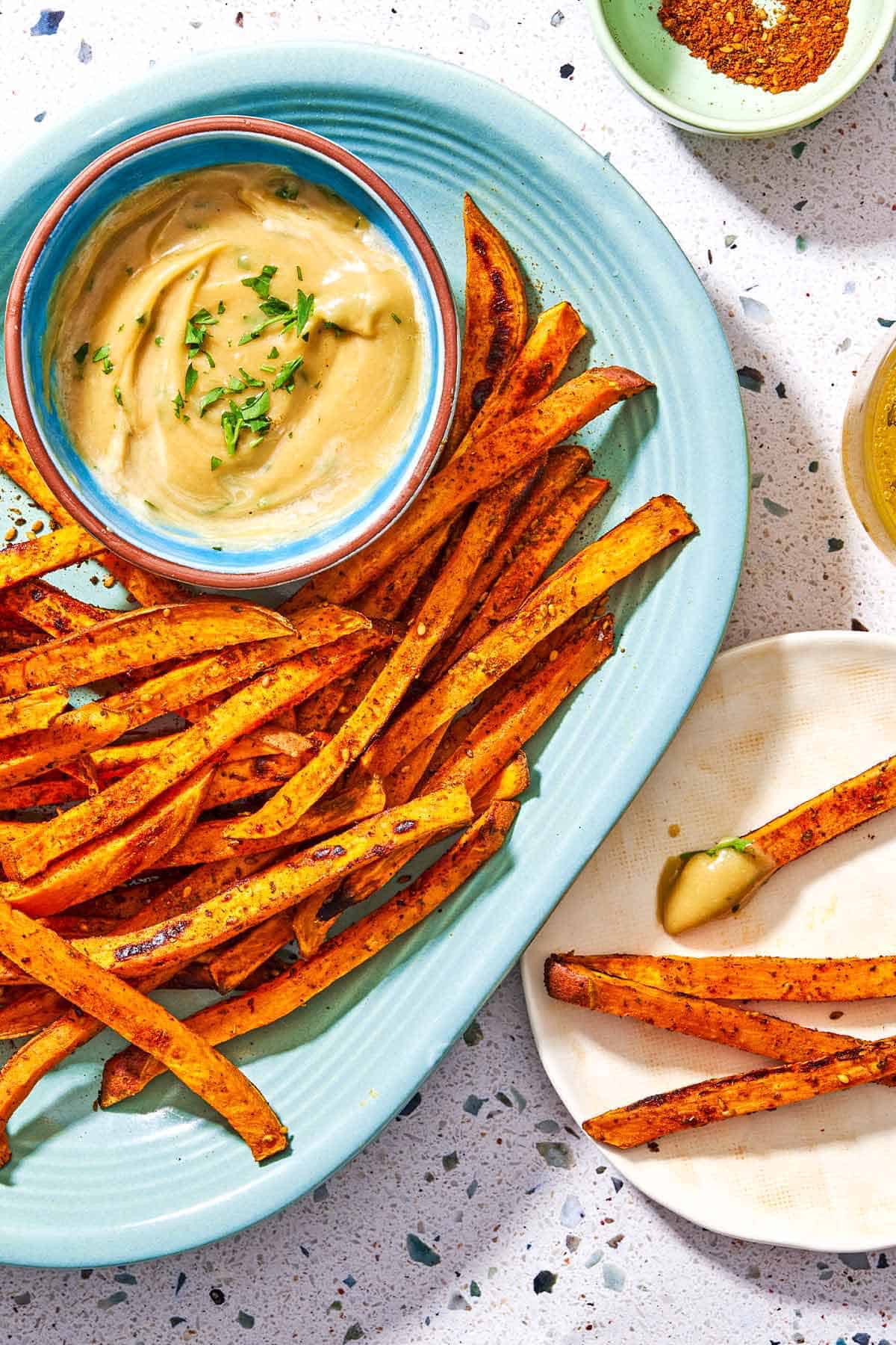 Sweet potato fries on a platter with a bowl of tahini sauce. Next to this is a bowl of the seasonings, and a plate with 3 sweet potato fries and a dollap of the tahini.