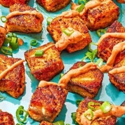 Several crispy salmon bites topped with a drizzle of harissa yogurt sauce and green onions on a plate with a juiced lemon half.