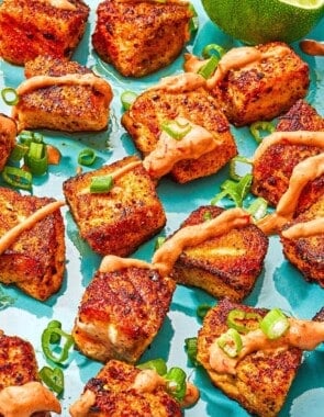 Several crispy salmon bites topped with a drizzle of harissa yogurt sauce and green onions on a plate with a juiced lemon half.