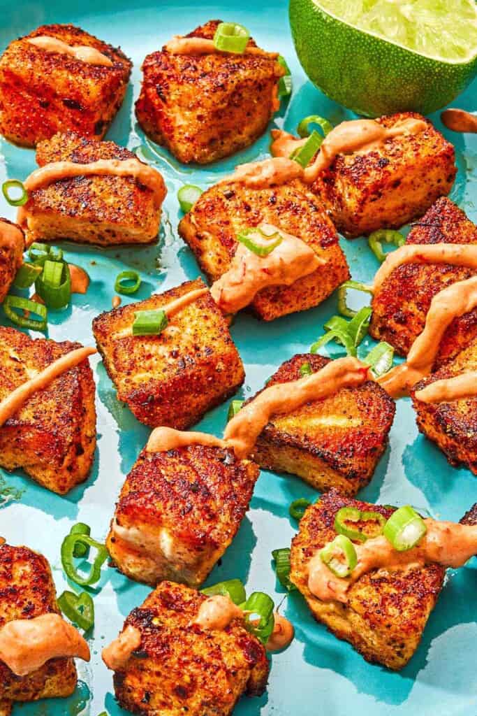 Several crispy salmon bites topped with a drizzle of harissa yogurt sauce and green onions on a plate with a juiced lemon half.