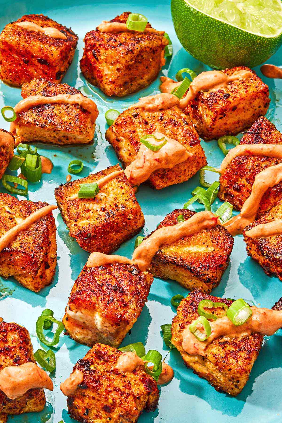 Several crispy salmon bites topped with a drizzle of harissa yogurt sauce and green onions on a plate with a juiced lemon half.