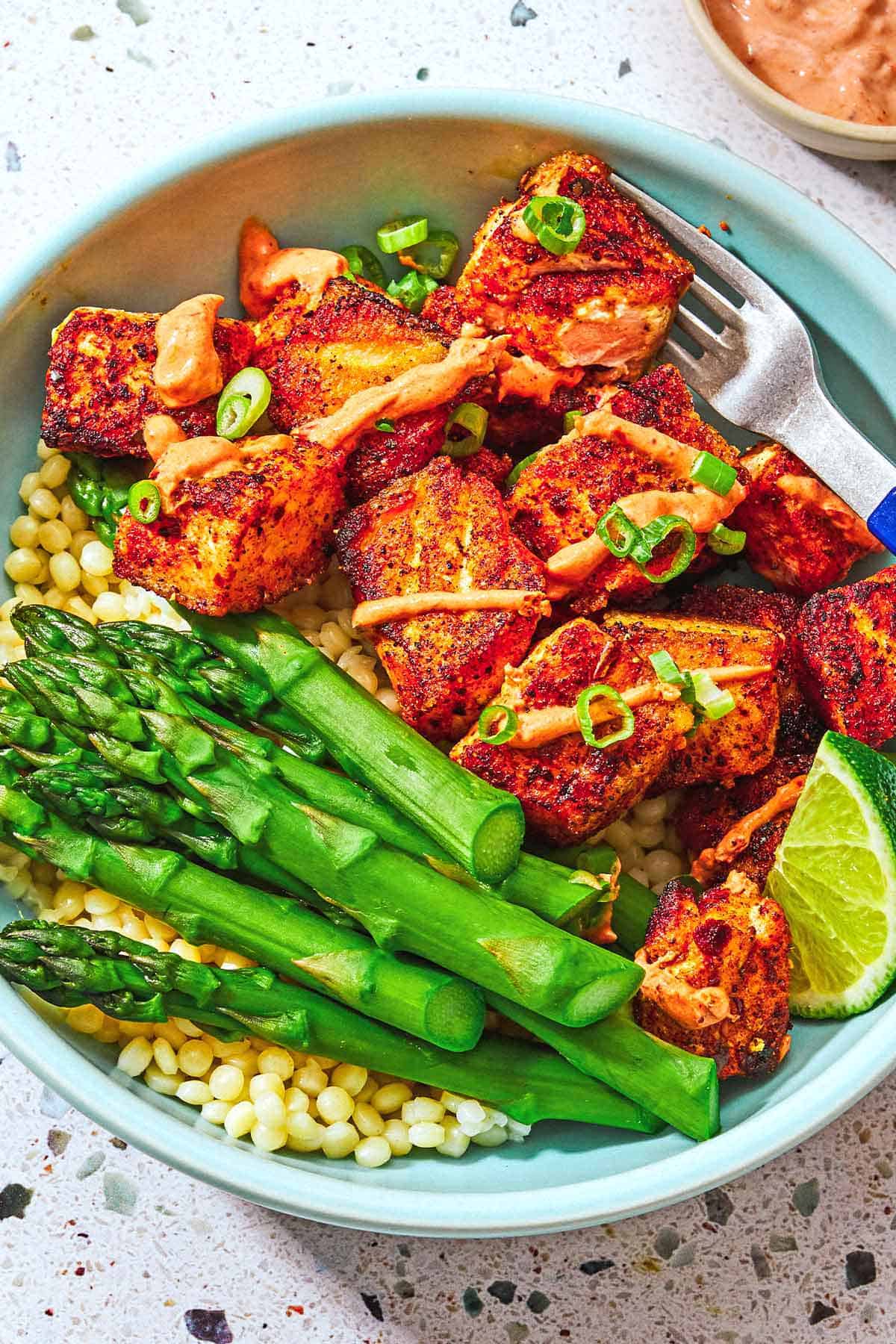 Several crispy salmon bites topped with a drizzle of harissa yogurt sauce and green onions and asparagus on a bed of pearl couscous in a bowl with a fork.