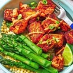 Several crispy salmon bites topped with a drizzle of harissa yogurt sauce and green onions and asparagus on a bed of pearl couscous in a bowl with a fork.