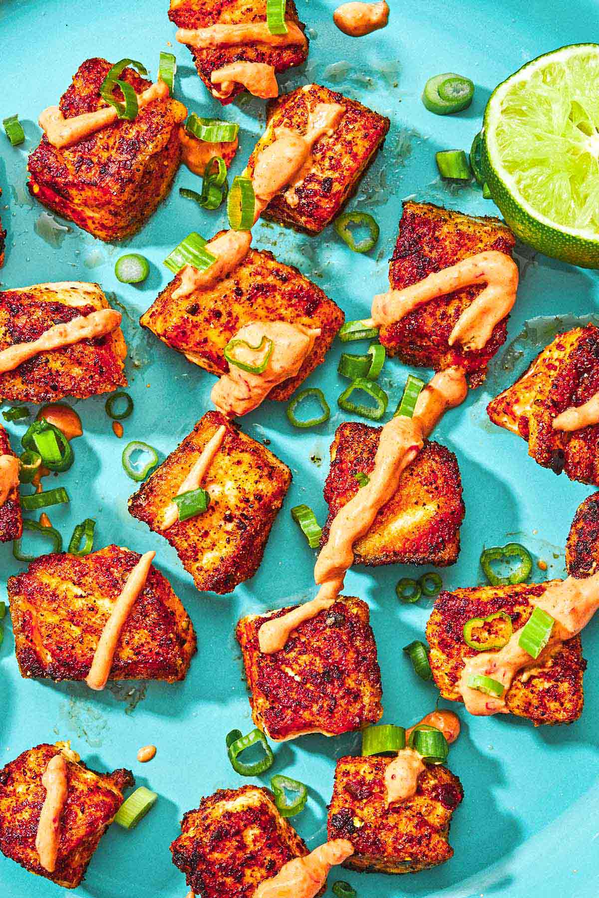 Several crispy salmon bites topped with a drizzle of harissa yogurt sauce and green onions on a plate with a juiced lemon half.
