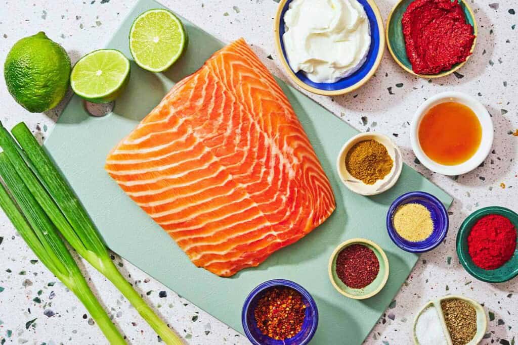 Ingredients for salmon bites including salmon, green onions, limes, greek yogurt, harissa paste, honey, smoked spanish paprika, garlic powder, aleppo flakes, cumin, sumac, salt, and black pepper.
