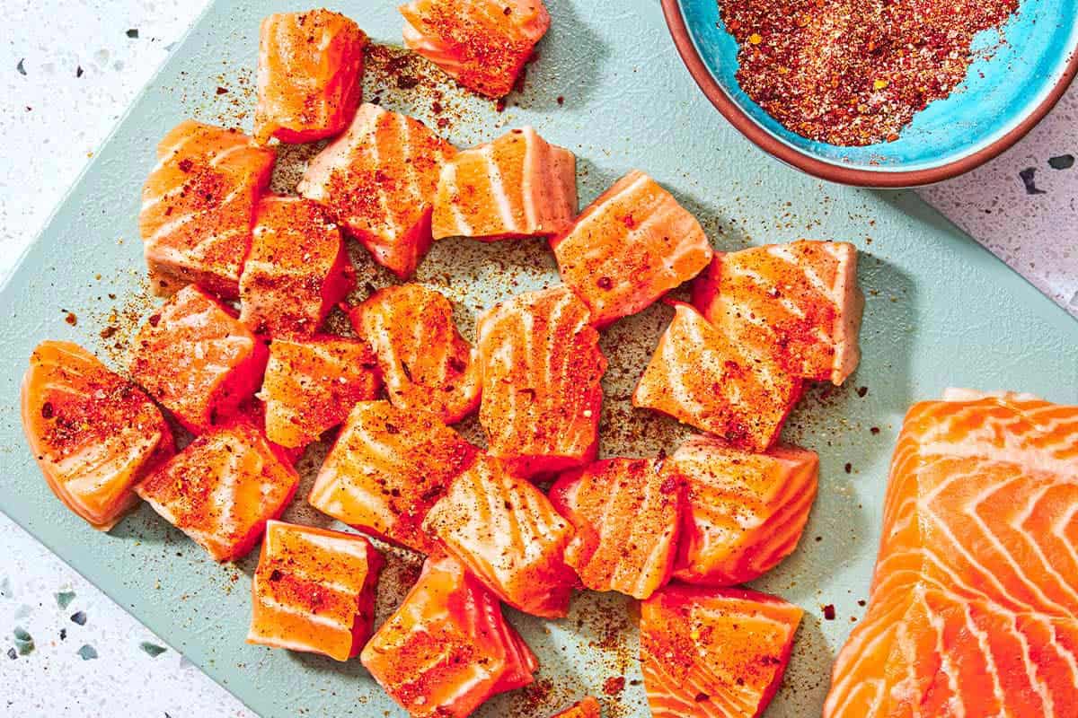 Uncooked seasoned salmon bites on a cutting board with the rest of the salmon fillet next to a bowl of the seasoning.