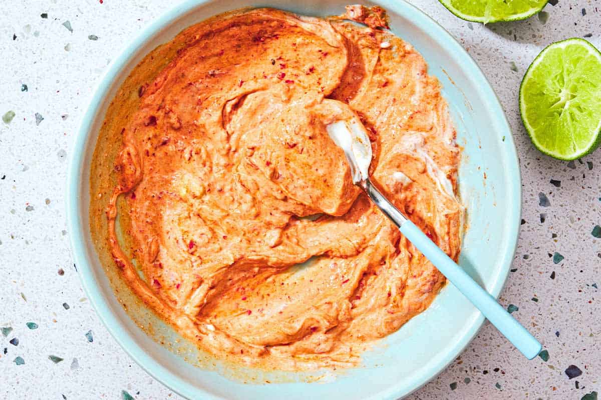 The harissa-yogurt sauce for the salmon bites in a bowl with a spoon next to two juiced lime halves.