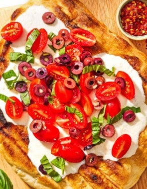 A grilled pizza on a wooden serving board with a bowl of red pepper flakes and some basil leaves.