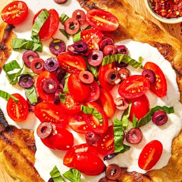 A grilled pizza on a wooden serving board with a bowl of red pepper flakes and some basil leaves.