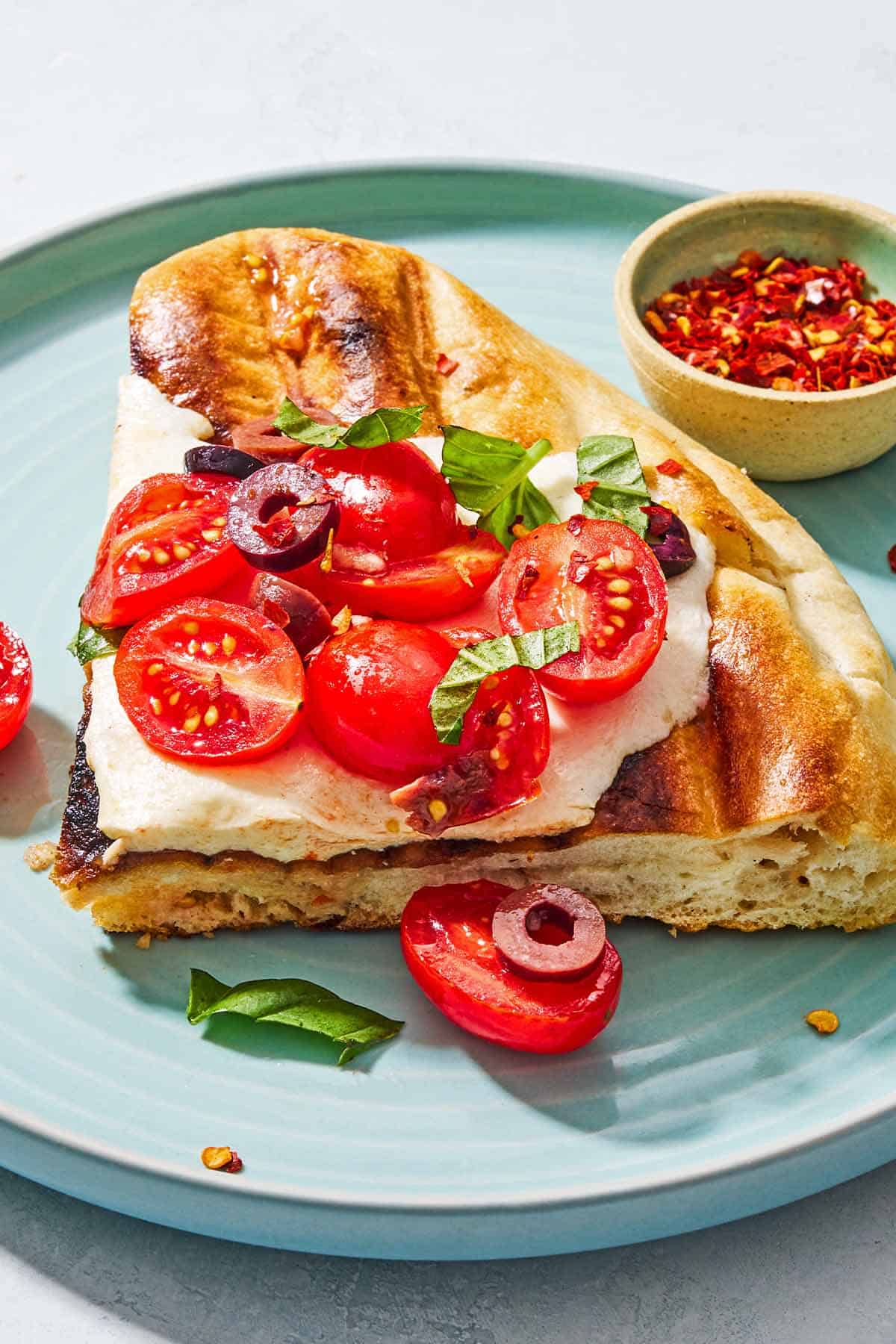 A slice of grilled pizza on a plate with a small bowl of red pepper flakes.