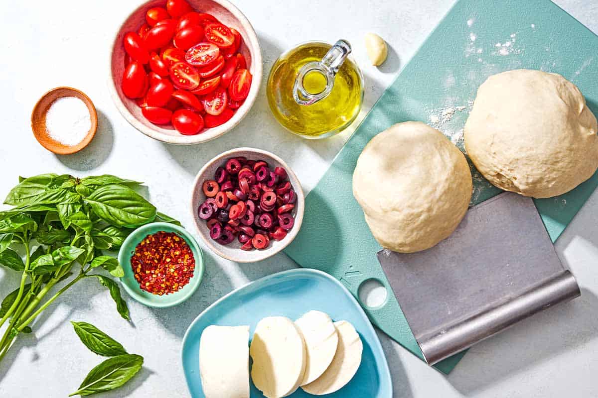 Ingredients for grilled pizza including 2 balls of dough, bowls sliced grape tomatoes, sliced kalamata olives, red pepper flakes, salt, a plate with mozzarella slices and fresh basil.