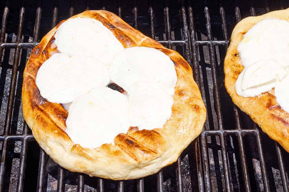The grilled pizza crust topped with slices of mozzarella cheese baking on a grill.