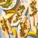Four trout fillets topped with the parsley caper sauce on a platter with lemon wedges, and a bowl of the parsley caper sauce with a spoon.