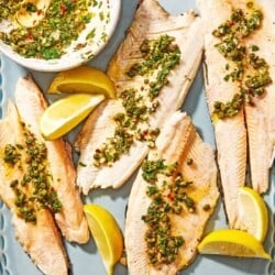 Four trout fillets topped with the parsley caper sauce on a platter with lemon wedges, and a bowl of the parsley caper sauce with a spoon.
