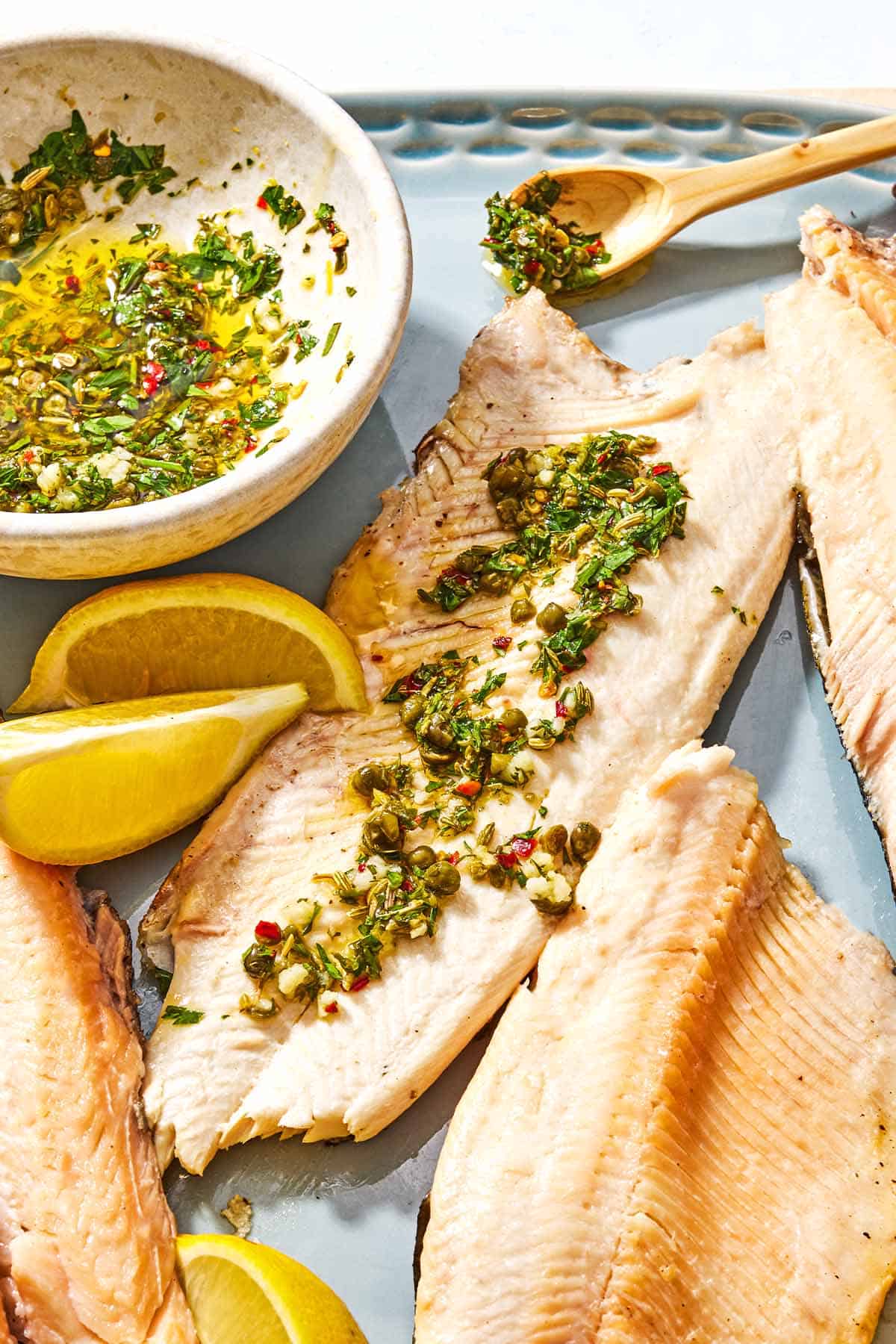 A trout fillet topped with parsley caper sauce on a platter with more fillets, lemon wedges, a spoon, and a bowl of the parsley caper sauce.