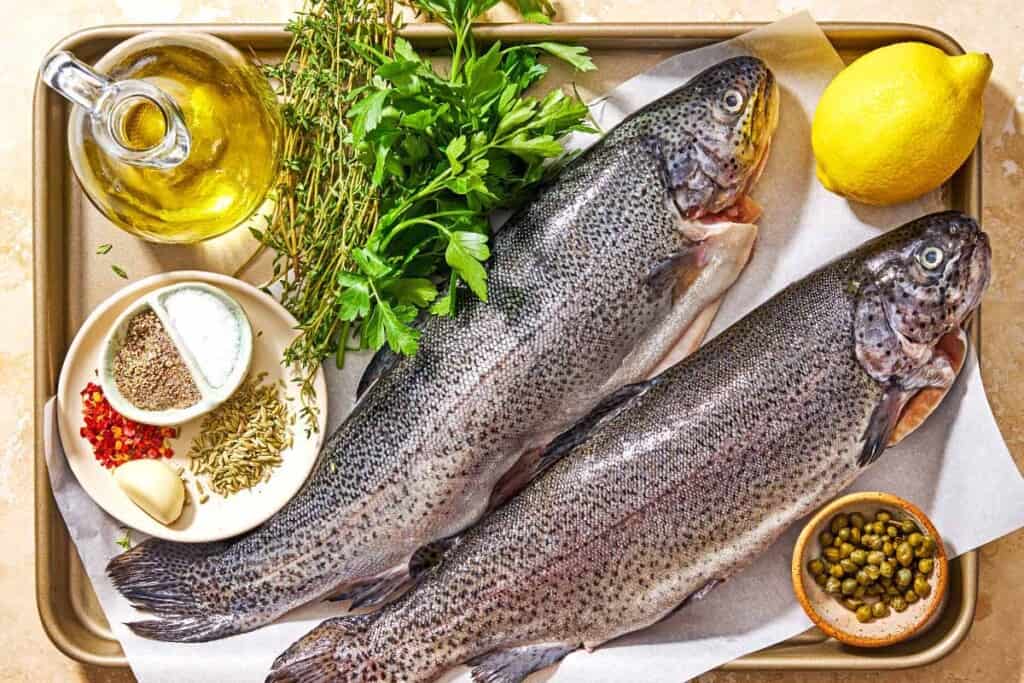 Ingredients for grilled trout including whole trout a lemon, capers, olive oil, salt, black pepper, parsley, thyme, garlic, red pepper flakes, and fennel seeds.