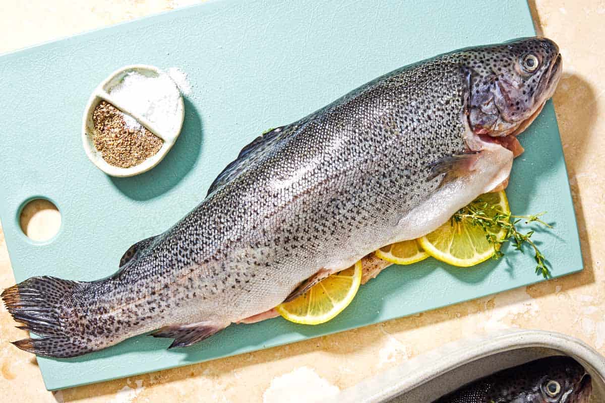 Two whole trout stuffed with lemon wheels and sprigs of thyme on a cutting board with a bowl of salt and black pepper.