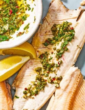 A trout fillet topped with parsley caper sauce on a platter with more fillets, lemon wedges, and a bowl of the parsley caper sauce.