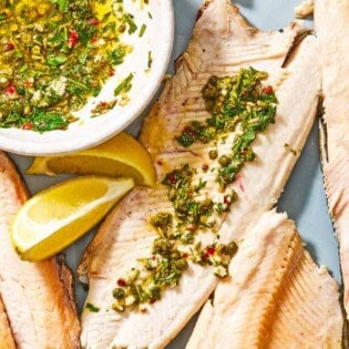 A trout fillet topped with parsley caper sauce on a platter with more fillets, lemon wedges, and a bowl of the parsley caper sauce.