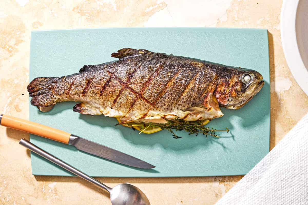 A whole grilled trout stuffed with lemon wheels and sprigs of thyme on a cutting board with a knife and spoon.