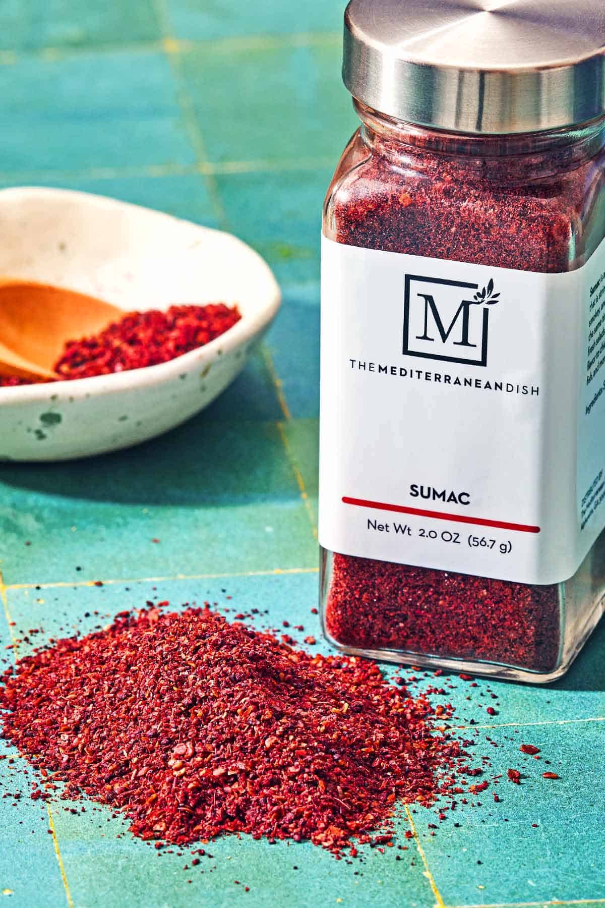 A bottle of sumac from the mediterranean dish next to a bowl or sumac with a spoon, and a handful of the sumac on the table.