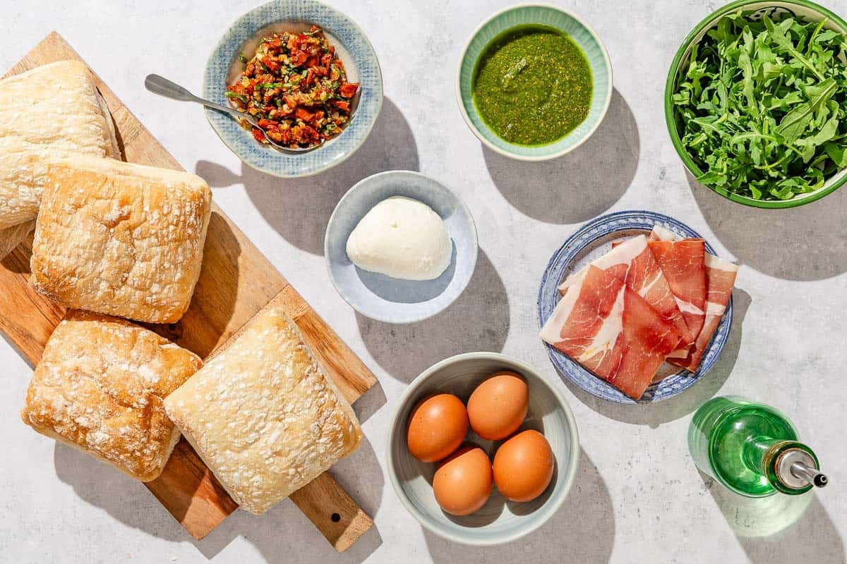 Ingredients for the breakfast sandwich recipe including eggs, mozzarella, pesto, prosciutto, arugula, olive oil and sun dried tomato spread.