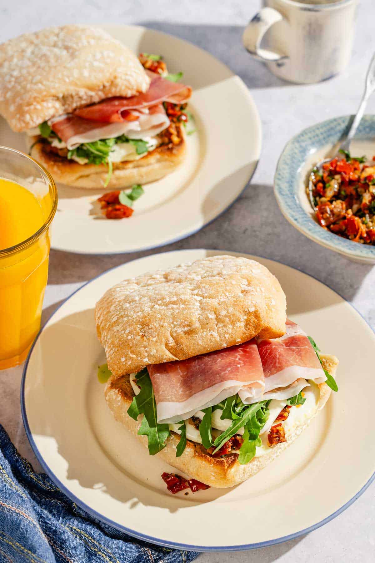 Two breakfast sandwiches on 2 plates next to a cloth napkin, a glass of orange juice, a bowl of sun dried tomato spread with a spoon, and a cup of coffee.