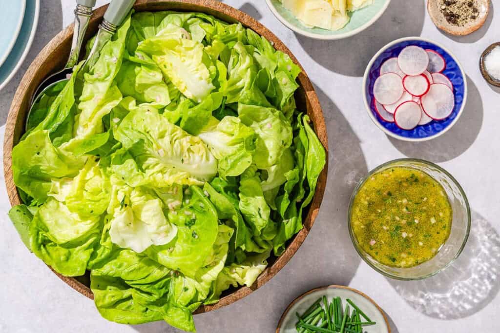 Butter lettuce salad in a serving bowl with serving utensils. Next to this is a stack of 2 plates, a plate of chives, and bowls of dressing, sliced radishes, shaved parmesan cheese, salt and black pepper.
