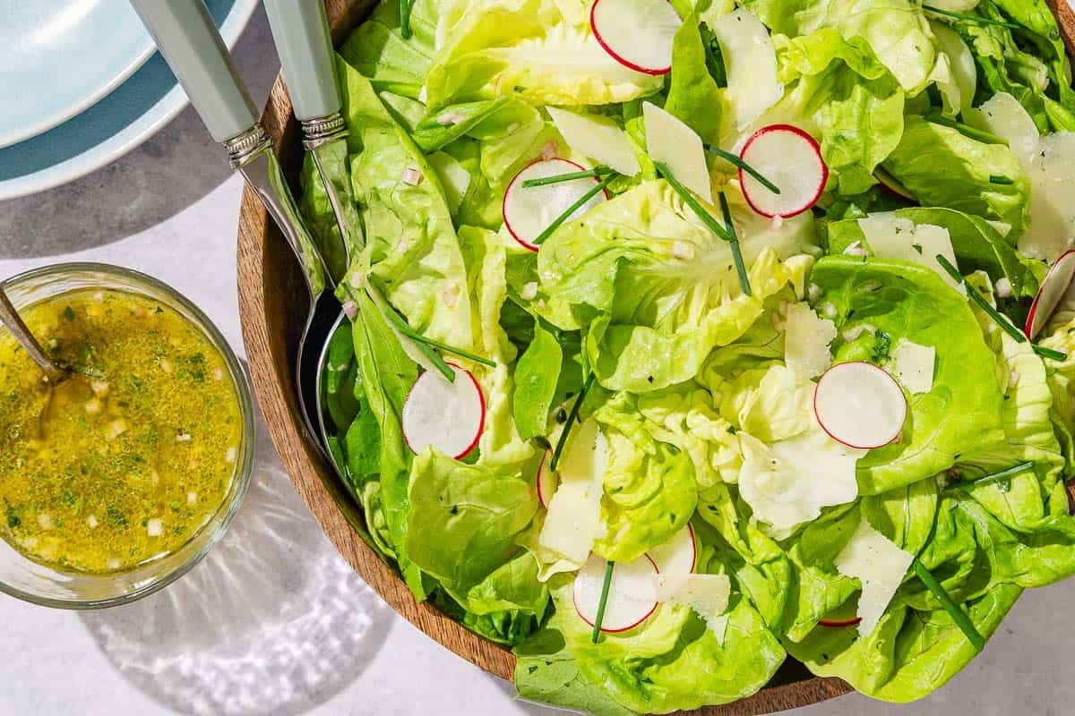 Butter lettuce salad in a serving bowl with serving utensils. Next to this is a stack of 2 plates and a bowl of dressing with a spoon.