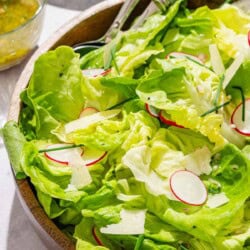 Butter lettuce salad in a serving bowl with serving utensils. Next to this is a stack of 2 plates and a bowl of dressing with a spoon.