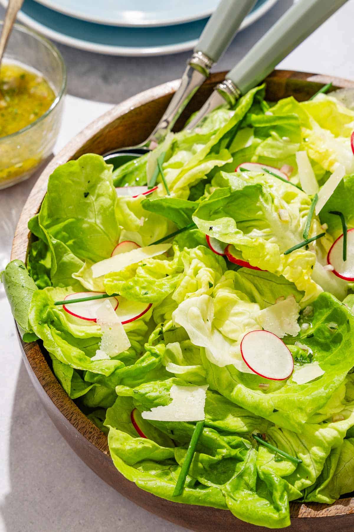 Butter lettuce salad in a serving bowl with serving utensils. Next to this is a stack of 2 plates and a bowl of dressing with a spoon.