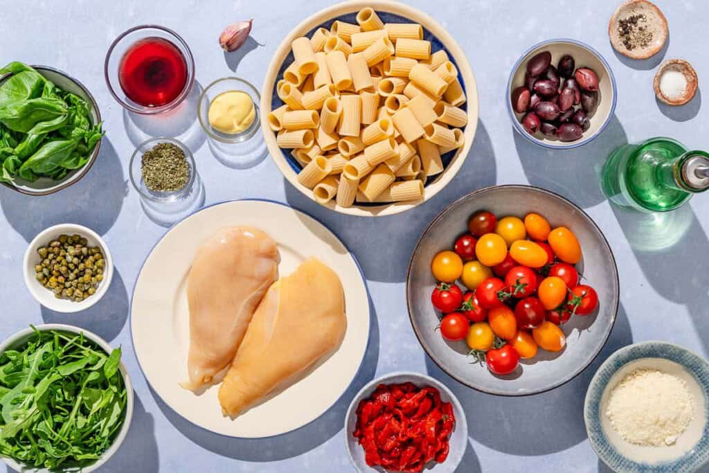 Ingredients for chicken pasta salad including chicken breasts, olive oil, Italian seasoning, salt, black pepper, rigatoni, grape tomatoes, roasted red peppers, kalamata olives, capers, arugula, basil, grated parmesan cheese, red wine vinegar, dijon mustard, and garlic.