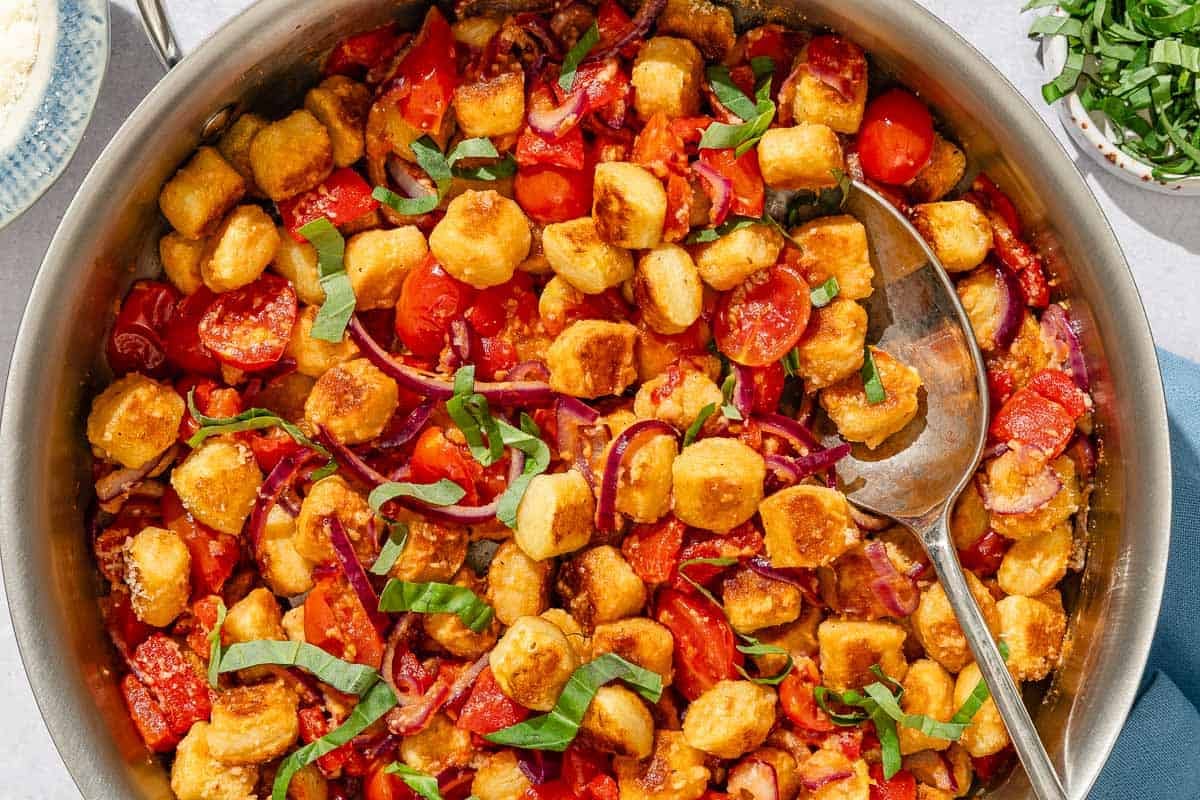Crispy gnocchi with cherry tomato sauce in a tall sided skillet with a serving spoon. Next to this is a small bowl of basil ribbons.