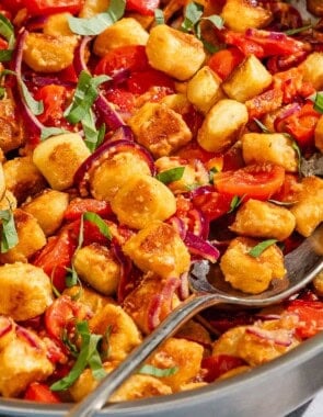 Crispy gnocchi with cherry tomato sauce in a tall sided skillet with a serving spoon. Next to this is a small bowl of basil ribbons.