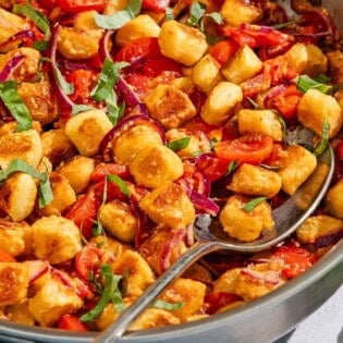 Crispy gnocchi with cherry tomato sauce in a tall sided skillet with a serving spoon. Next to this is a small bowl of basil ribbons.