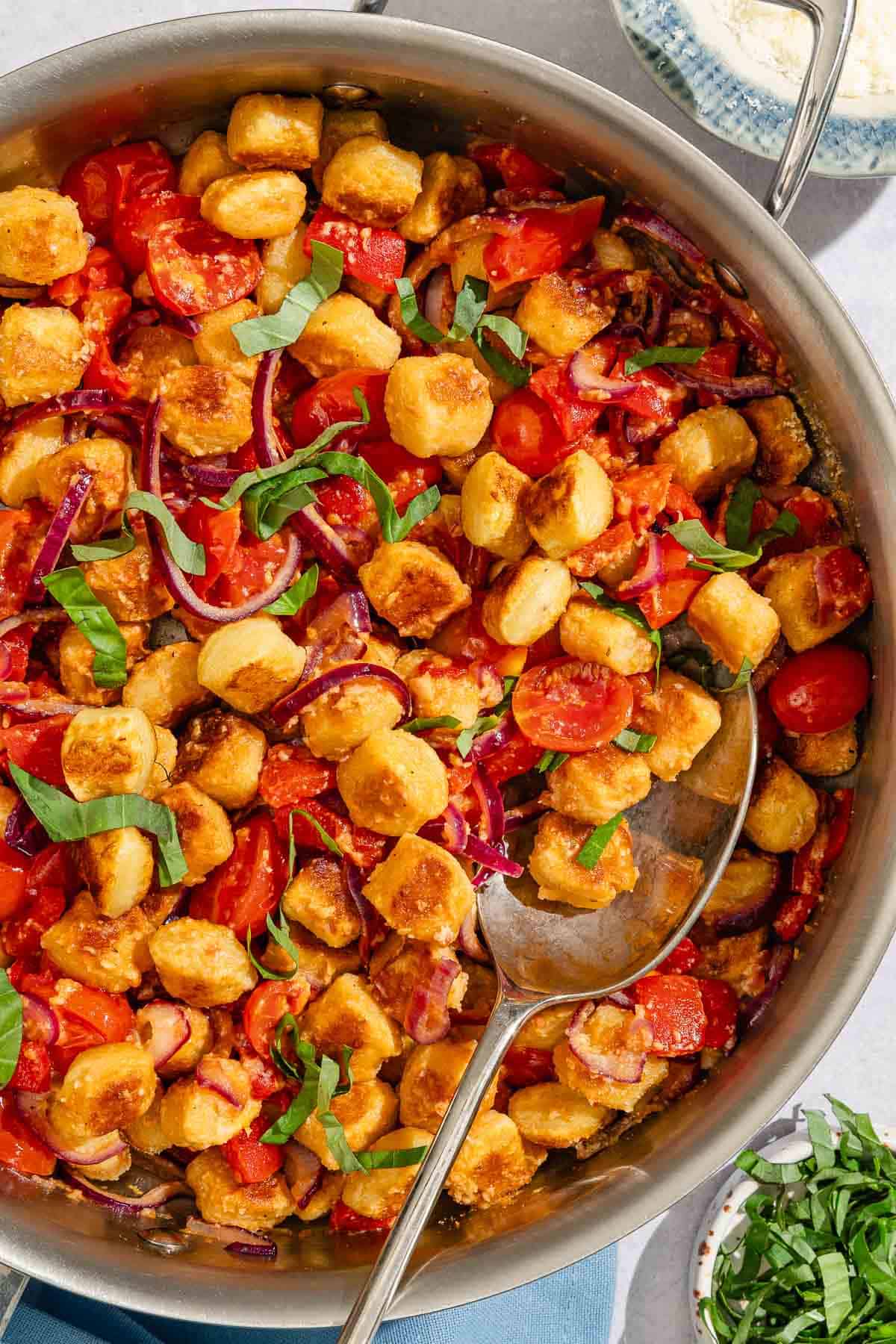 Crispy gnocchi with cherry tomato sauce in a tall sided skillet with a serving spoon. Next to this is a small bowl of basil ribbons.