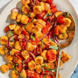 Crispy gnocchi with cherry tomato sauce on a platter with a serving spoon. Next to this is a small bowl of basil ribbons and a stack of 2 plates.