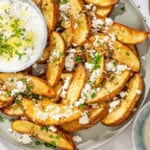 Greek fries topped with parsley and crumbled feta on a platter with a bowl of tzatziki sauce next to bowls of chopped parsley and crumbled feta.