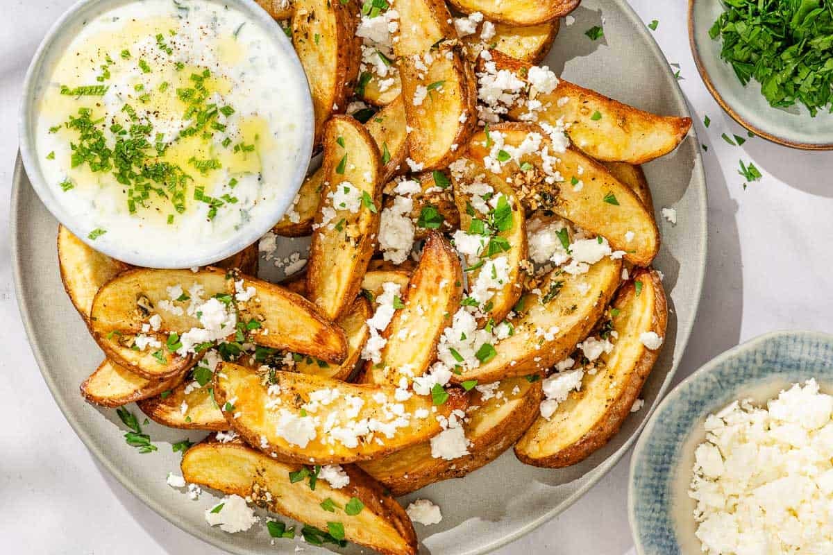 Greek fries topped with parsley and crumbled feta on a platter with a bowl of tzatziki sauce next to bowls of chopped parsley and crumbled feta.