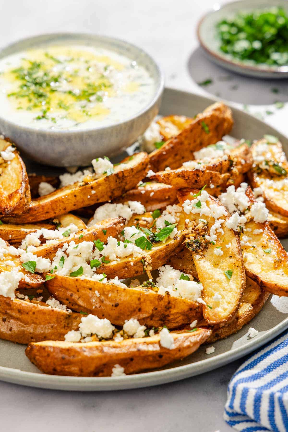 Greek fries topped with parsley and crumbled feta on a platter with a bowl of tzatziki sauce.