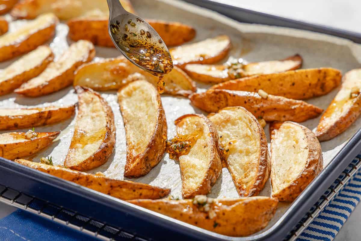 Baked potato wedges on a parchment lined sheet pan being drizzled with the sauce.