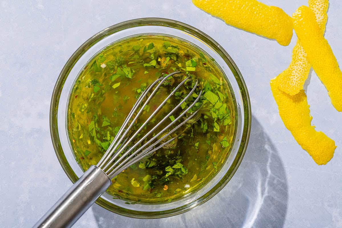 The dressing for the seafood salad in a bowl with a whisk next to some lemon peel.