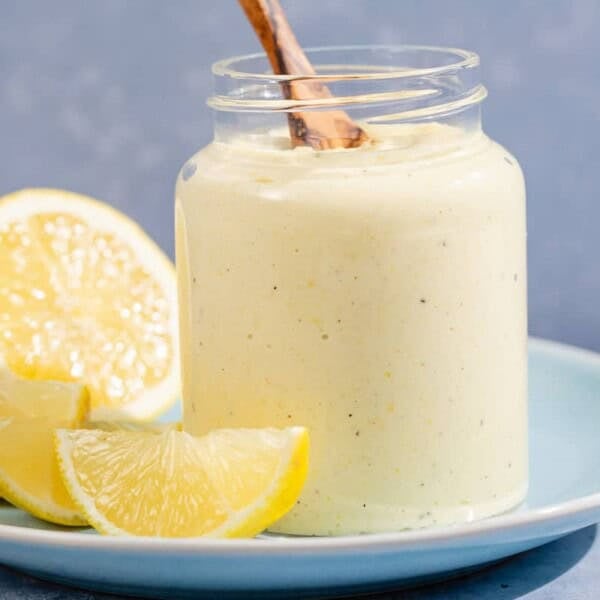 A jar of lemon pesto with a wooden spoon resting on a plate with lemon slices.