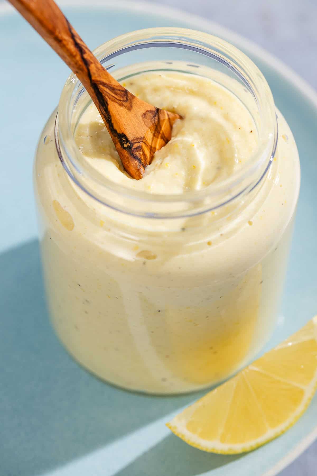 A jar of lemon pesto with a wooden spoon resting on a plate with a lemon slice.