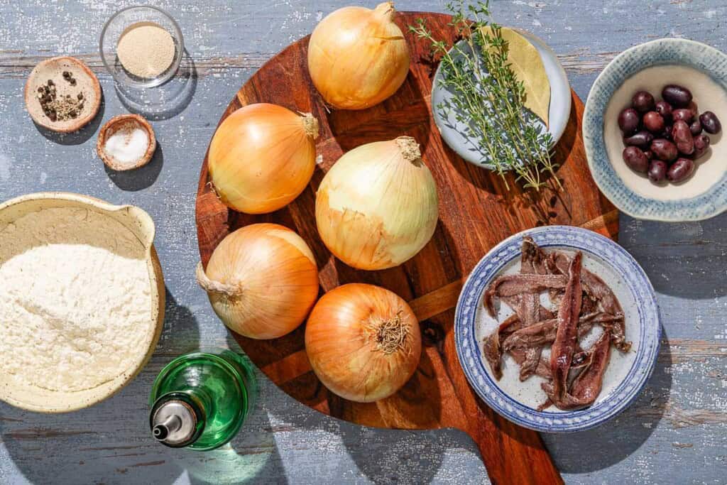 Ingredients for Pissaladière including flour, yeast, salt, olive oil, yellow onions, fresh thyme, a bay leaf, anchovy filets, and kalamata olives.